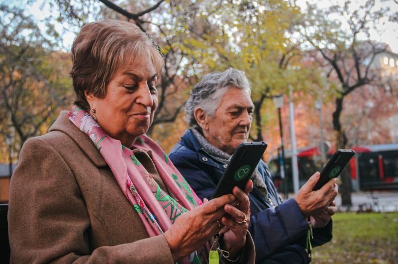 Imagen: dos mujeres mayores sentadas en un banco de un parque utilizan un móvil con el logo de Maximiliana.